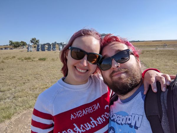 Cassey and a person with a beard and short pink hair, both wearing sunglasses and hugging, stand in the foreground with Carhenge in the background
