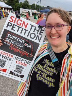 Cassey wears a shirt reading 'Nebraska Family Alliance is a Hate Group' and a rainbow button-up, and holds a petition for Support Our Schools Nebraska
